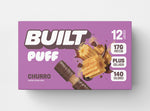 A purple box of 12 BUILT churro puffs on a white background.